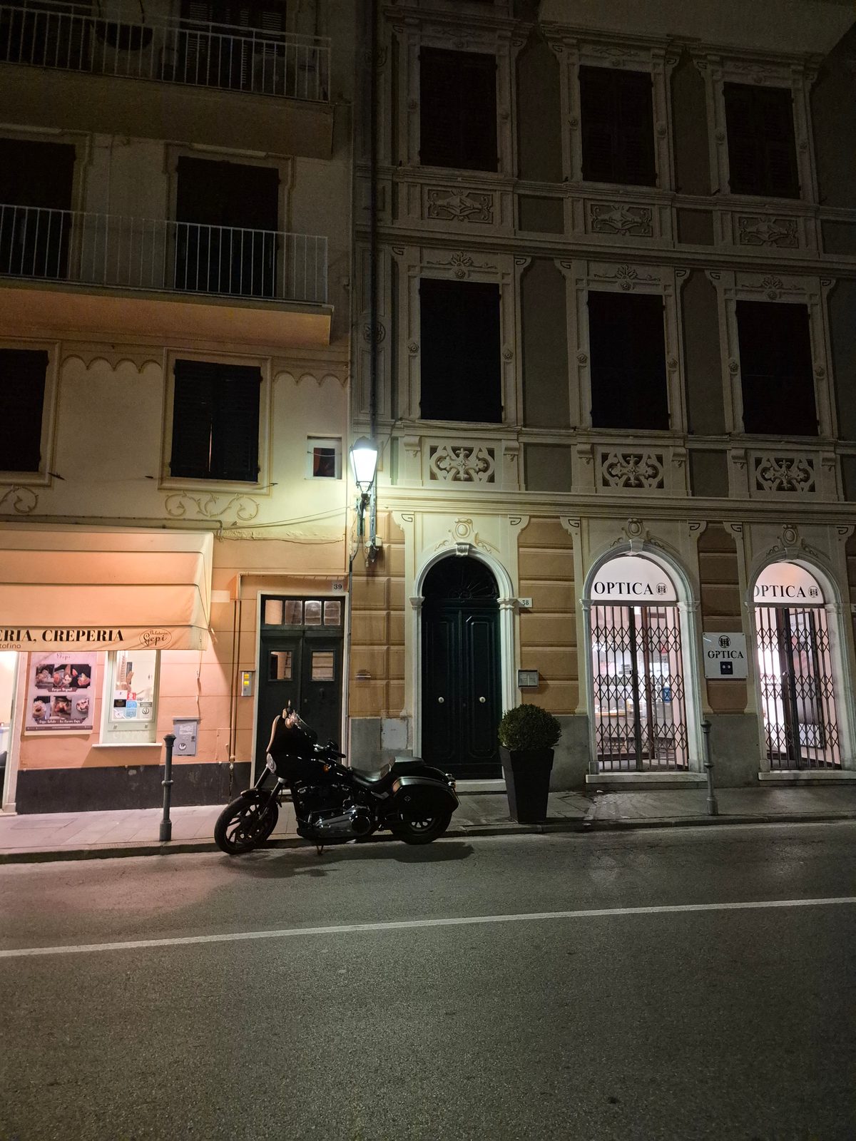 A black cruiser motorcycle parked at night on a street lined with old Italian buildings, with a creperia and an opticians shop.