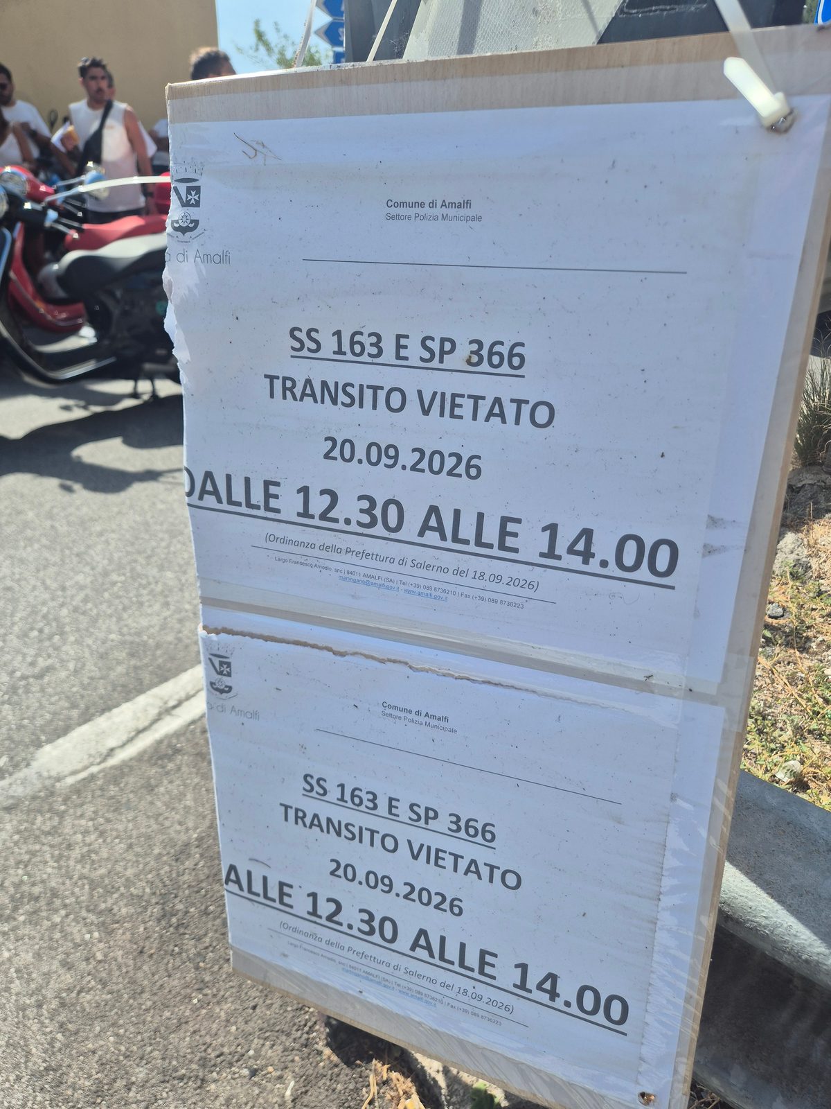 A roadside notice from the Comune di Amalfi closing the SS 163 and SP 366 to traffic between 12.30 and 14.00, with a red scooter parked behind.