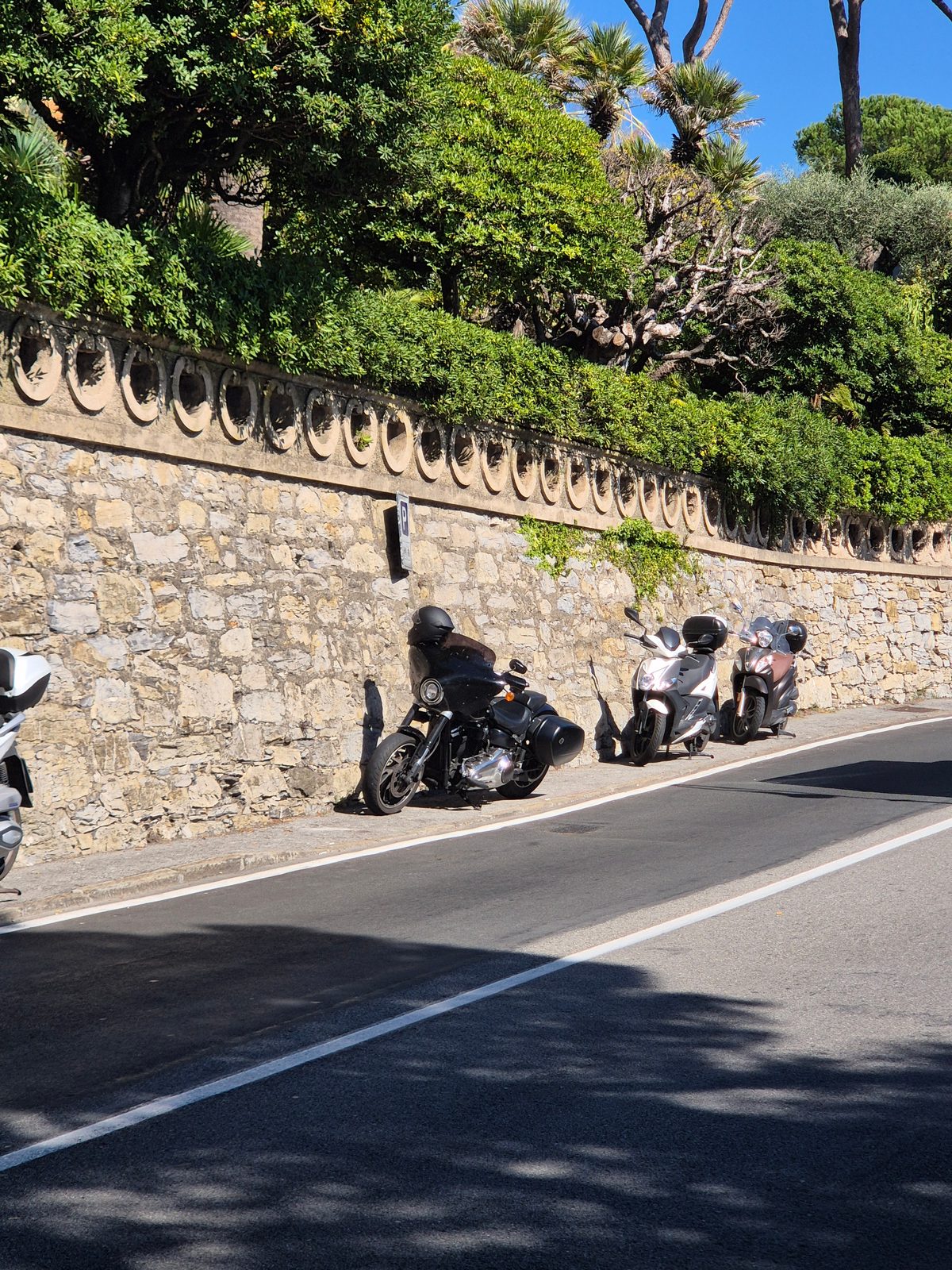A black motorcycle and two scooters parked beside an ornate stone wall on a sunny roadside.