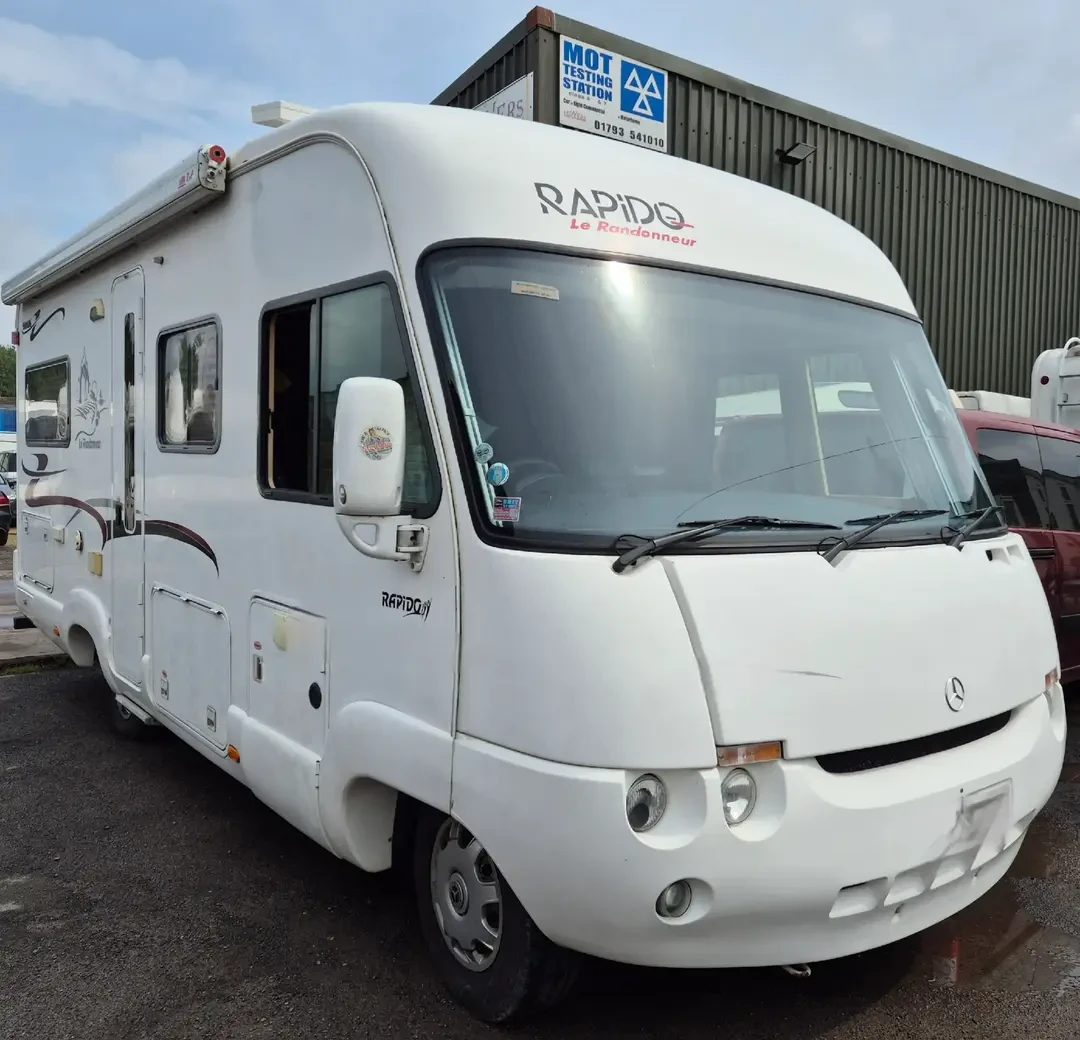 A white Rapido Le Randonneur A-class motorhome on a Mercedes base, parked outside a garage with an MOT Testing Station sign.