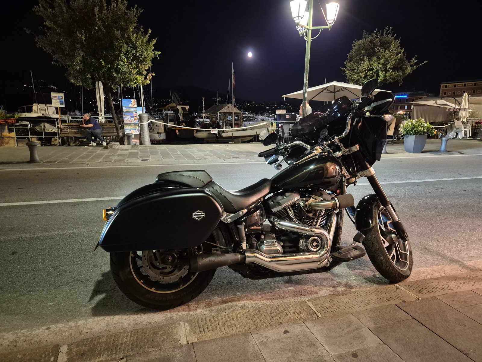A black Harley-Davidson motorcycle parked by a harbour at night with boats and a full moon in the background.