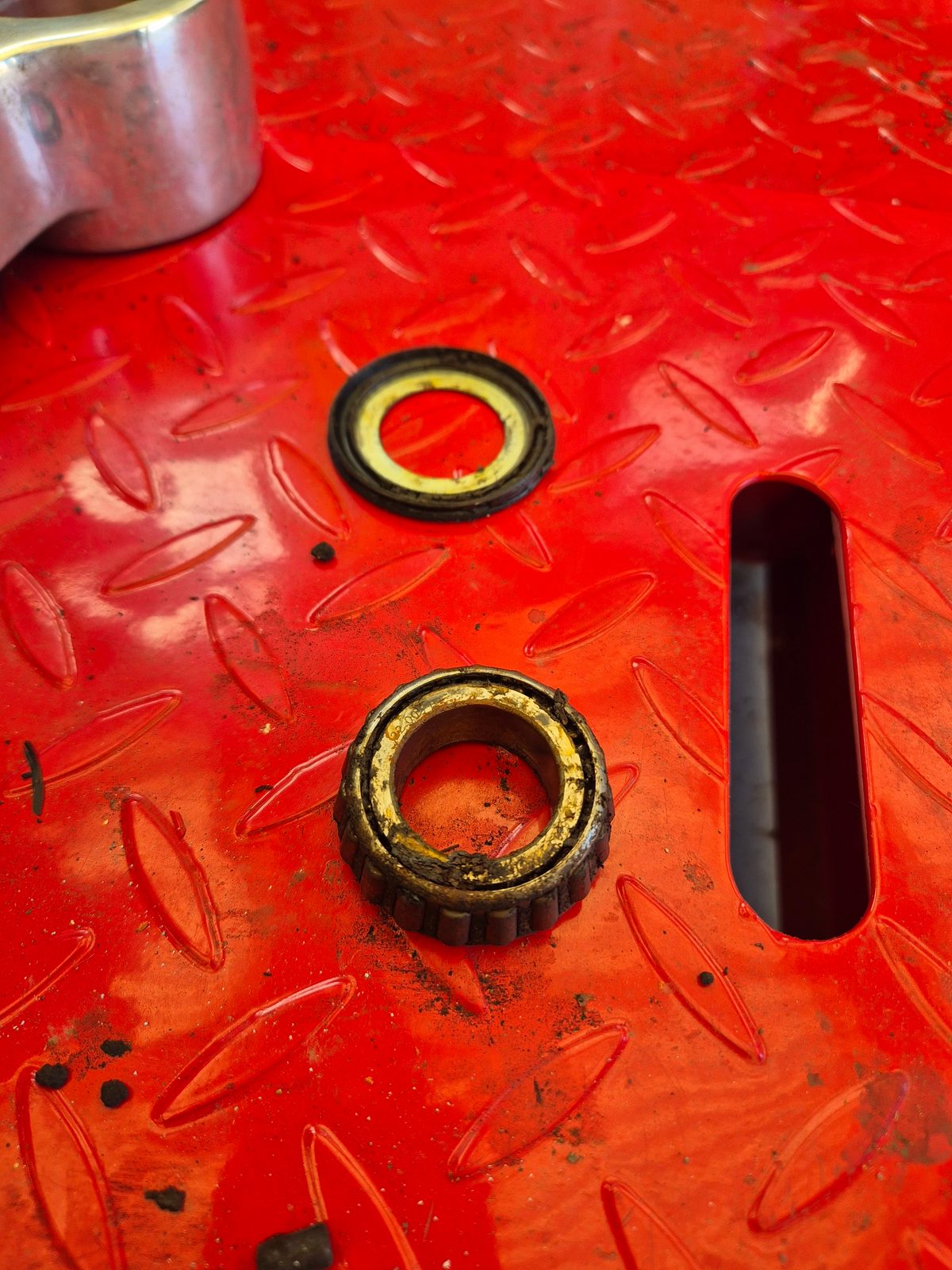 A worn tapered roller wheel bearing and a rubber oil seal resting on a red diamond-plate metal surface, with a metal socket in the corner.