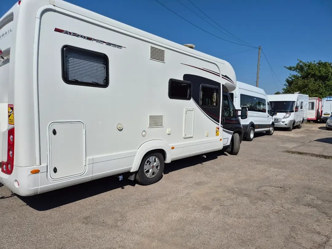 A white Autotrail Apache coachbuilt motorhome parked on tarmac with a minibus and further motorhomes behind under a clear blue sky.