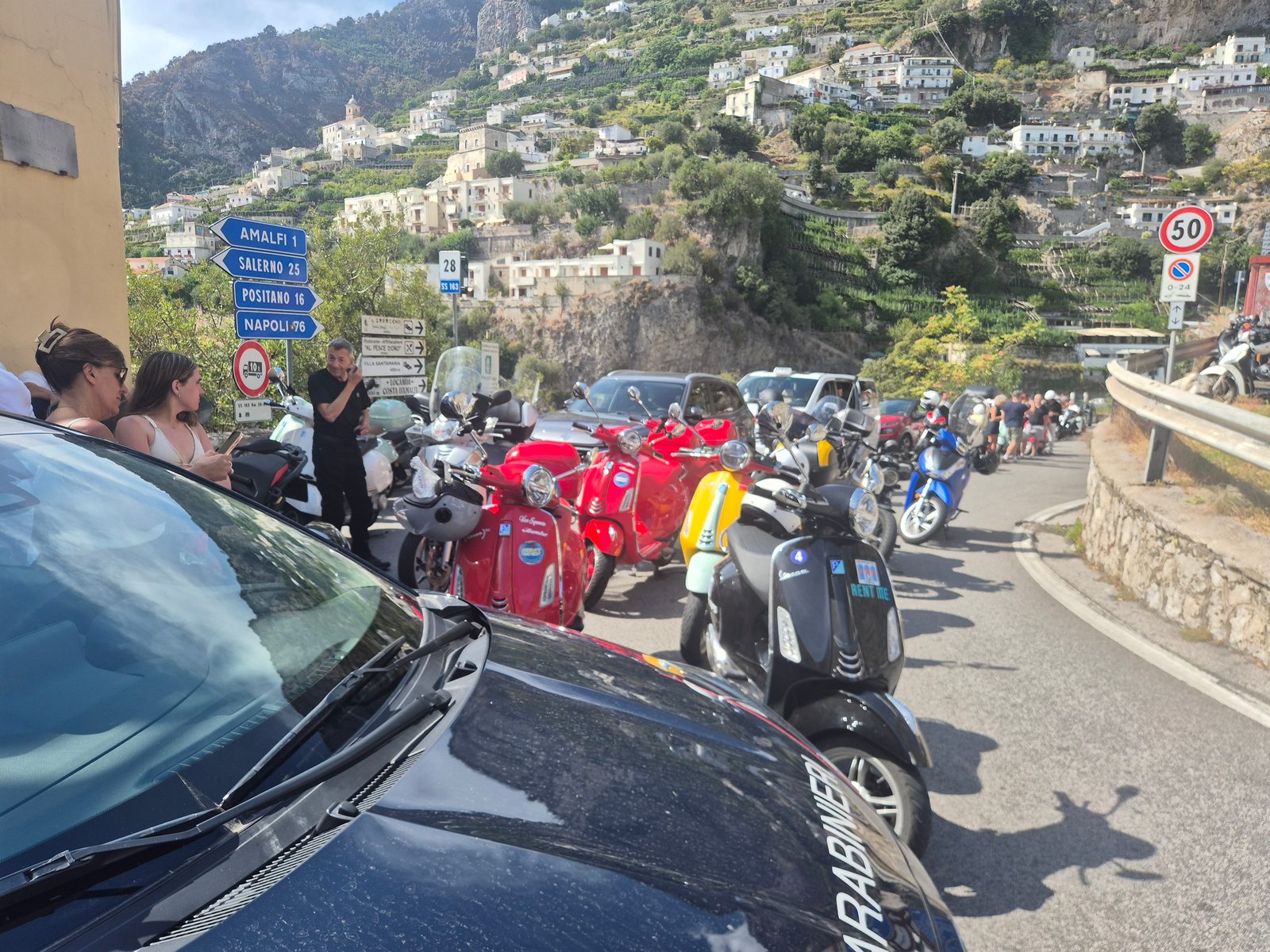 A row of parked scooters and Vespas on a busy road in Amalfi, Italy, with a hillside town and road signs in the background.