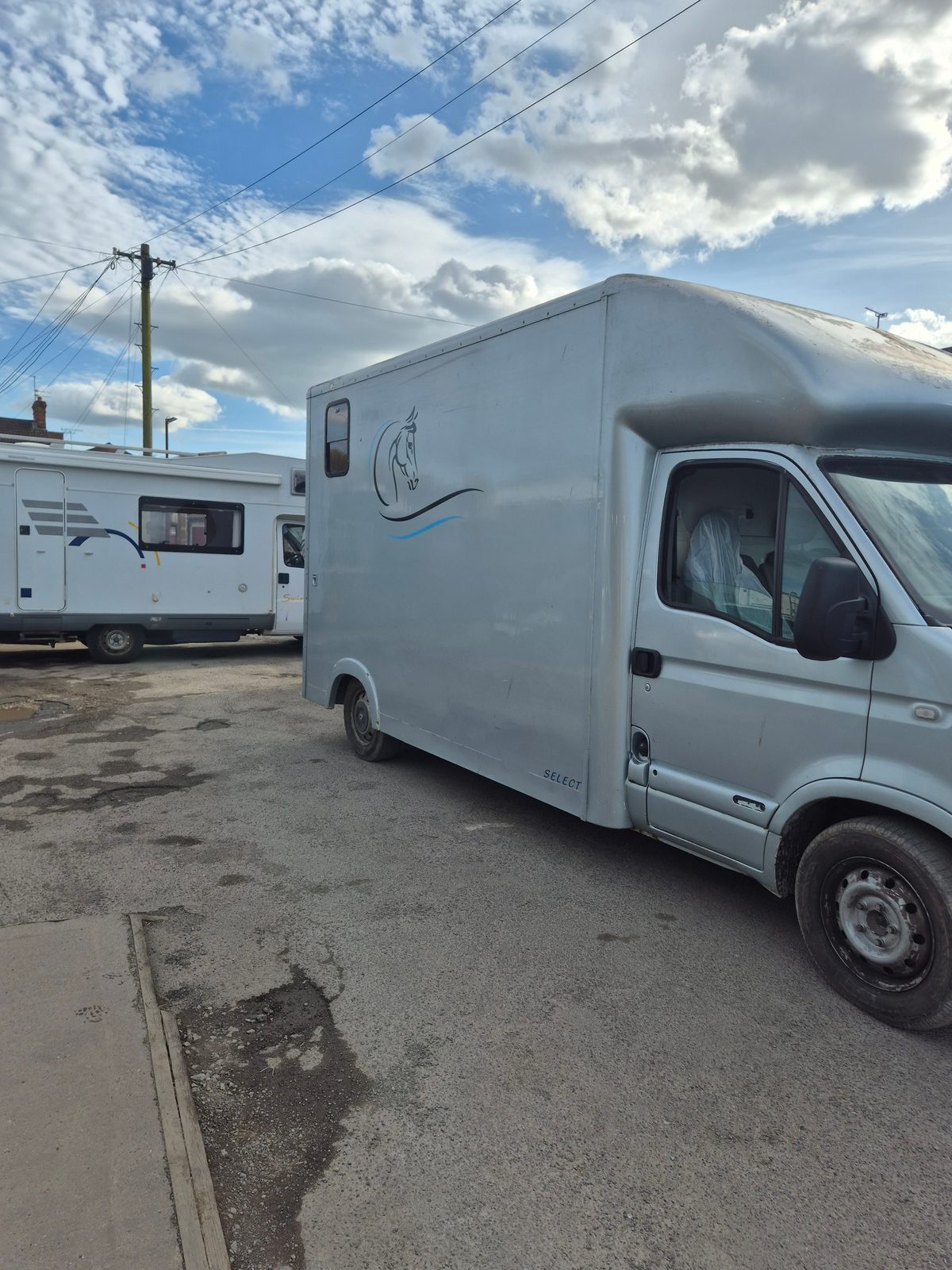 A pale blue coachbuilt horsebox van with a horse's head graphic, parked on a tarmac forecourt beside a white motorhome under a cloudy sky.