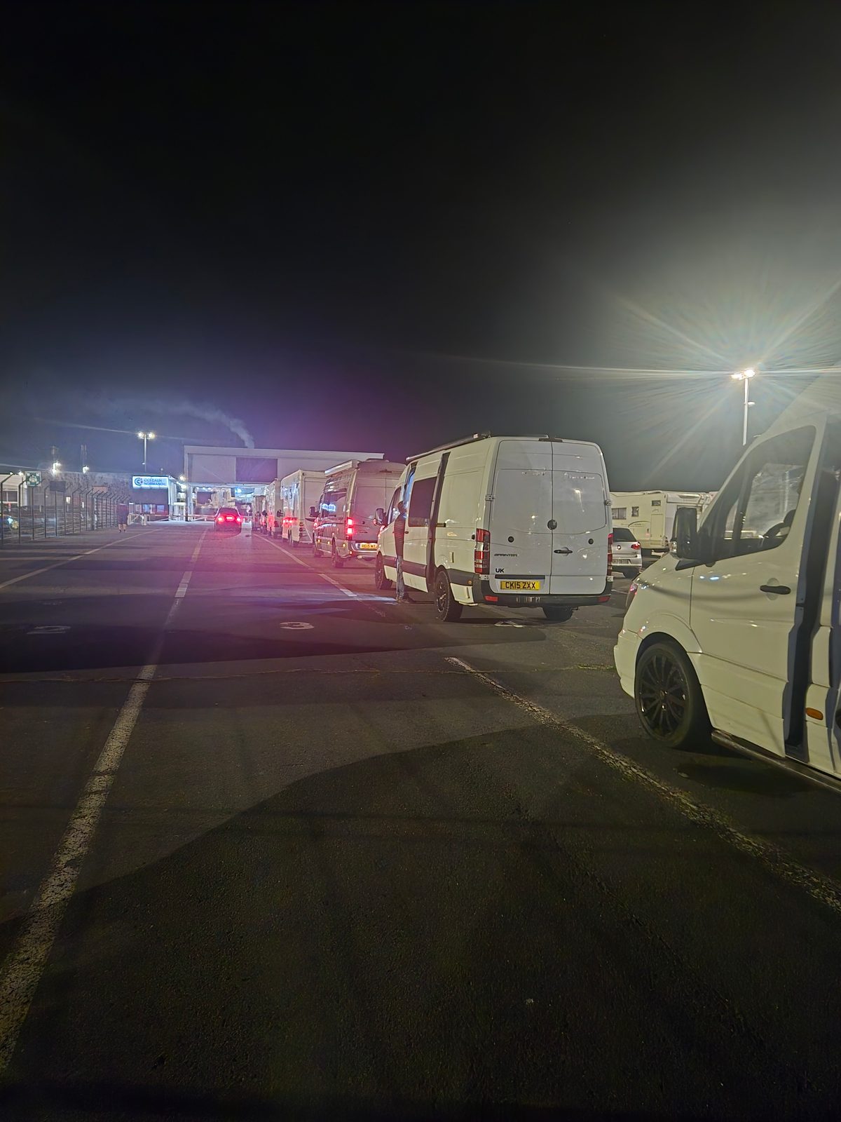 A line of vans queuing at night at a ferry port terminal under floodlights.