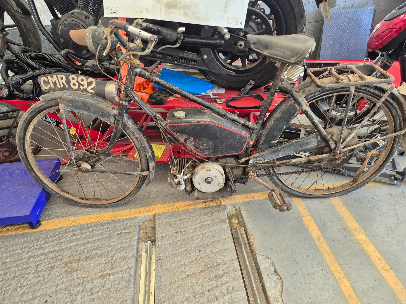 A dusty old black Norman autocycle with registration CMR 892 standing in the workshop, with a modern motorcycle on a ramp behind.