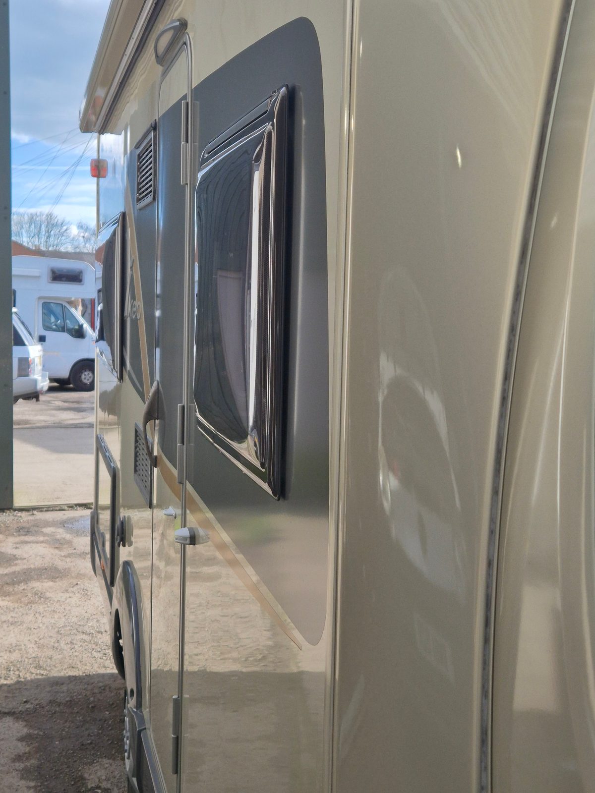 Close-up of a cream coachbuilt motorhome's side showing a service access door, hinged panel and ventilation grilles, with another motorhome parked in the background.