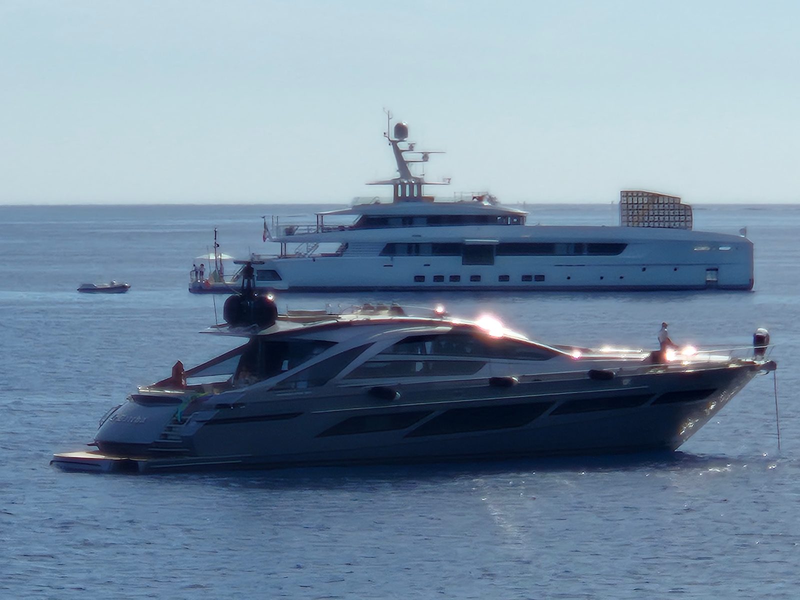 A sleek grey motor yacht anchored in front of a larger superyacht with a helicopter on deck, out at sea.